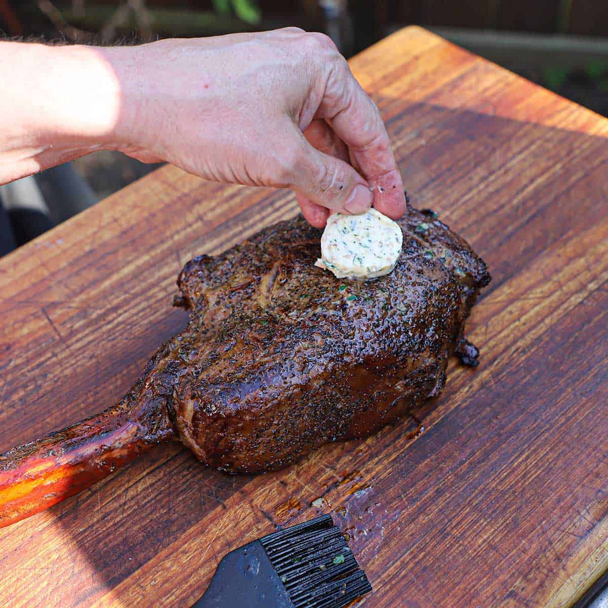 A person placing a chilled medallion of roasted garlic and bone marrow butter on top of a tomahawk steak that has just been reverse seared.