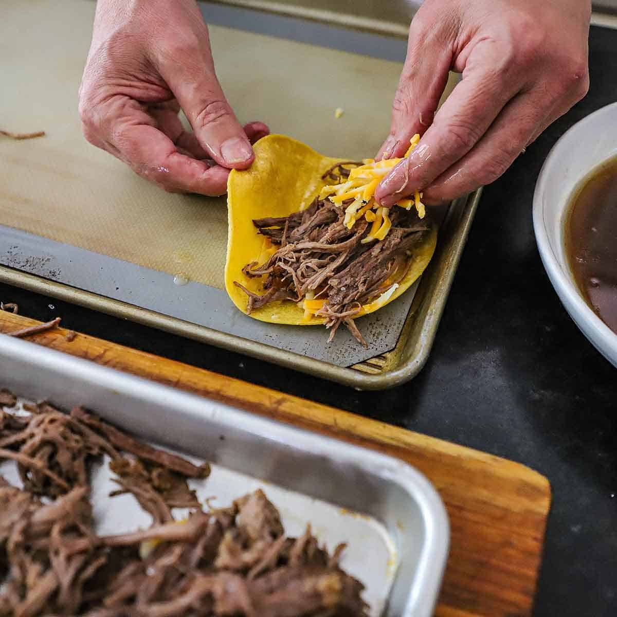 A person placing shredded cheese over a pile of shredded brisket in a lightly oiled corn tortilla on a baking sheet lined with a silicone matt.