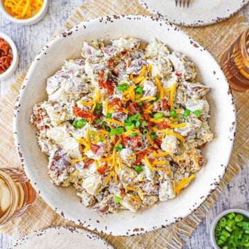 An overhead view of a white serving bowl filled with a creamy loaded potato salad topped with crispy bacon bits, chopped scallions, and shredded cheddar cheese.