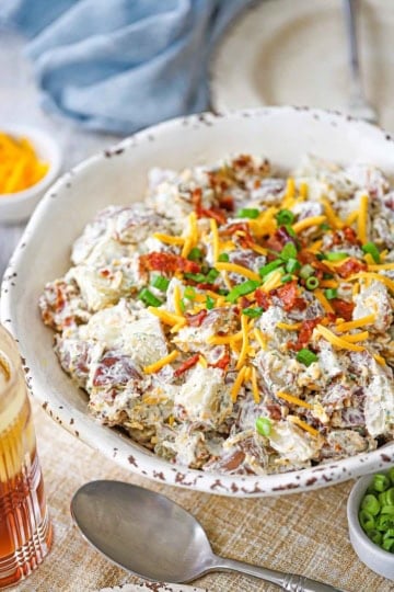 A side-view of a white serving bowl filled with a homemade creamy loaded potato salad topped with crispy bacon bits, chopped scallions, and shredded cheddar cheese.