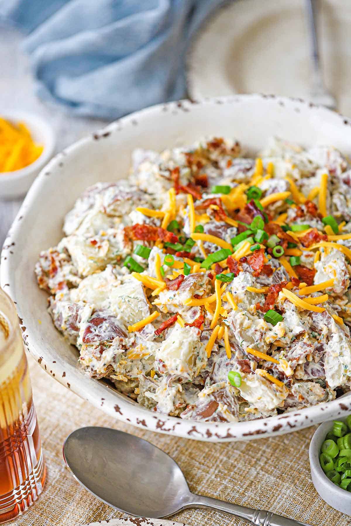 A side-view of a white serving bowl filled with a homemade creamy loaded potato salad topped with crispy bacon bits, chopped scallions, and shredded cheddar cheese.