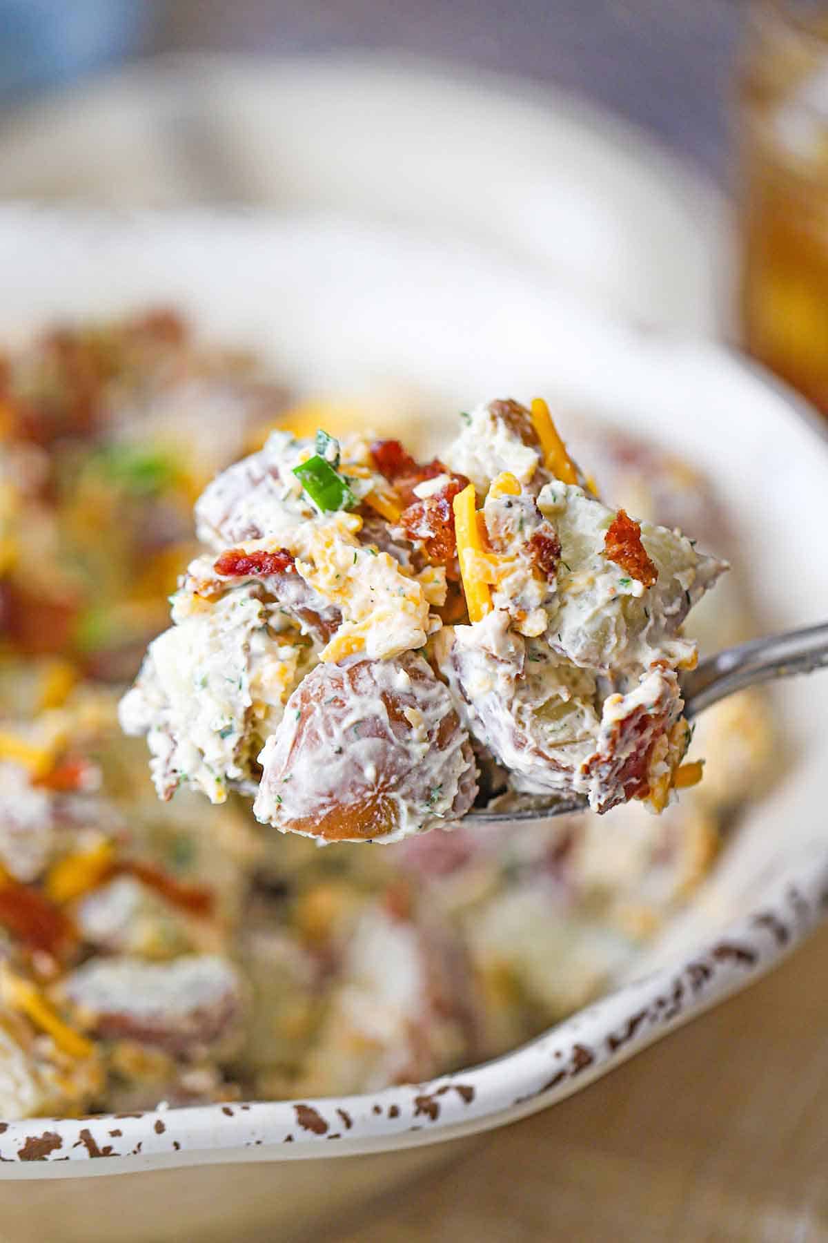 A large serving spoon holding a heaping mound of creamy loaded potato salad over a bowl of the same.