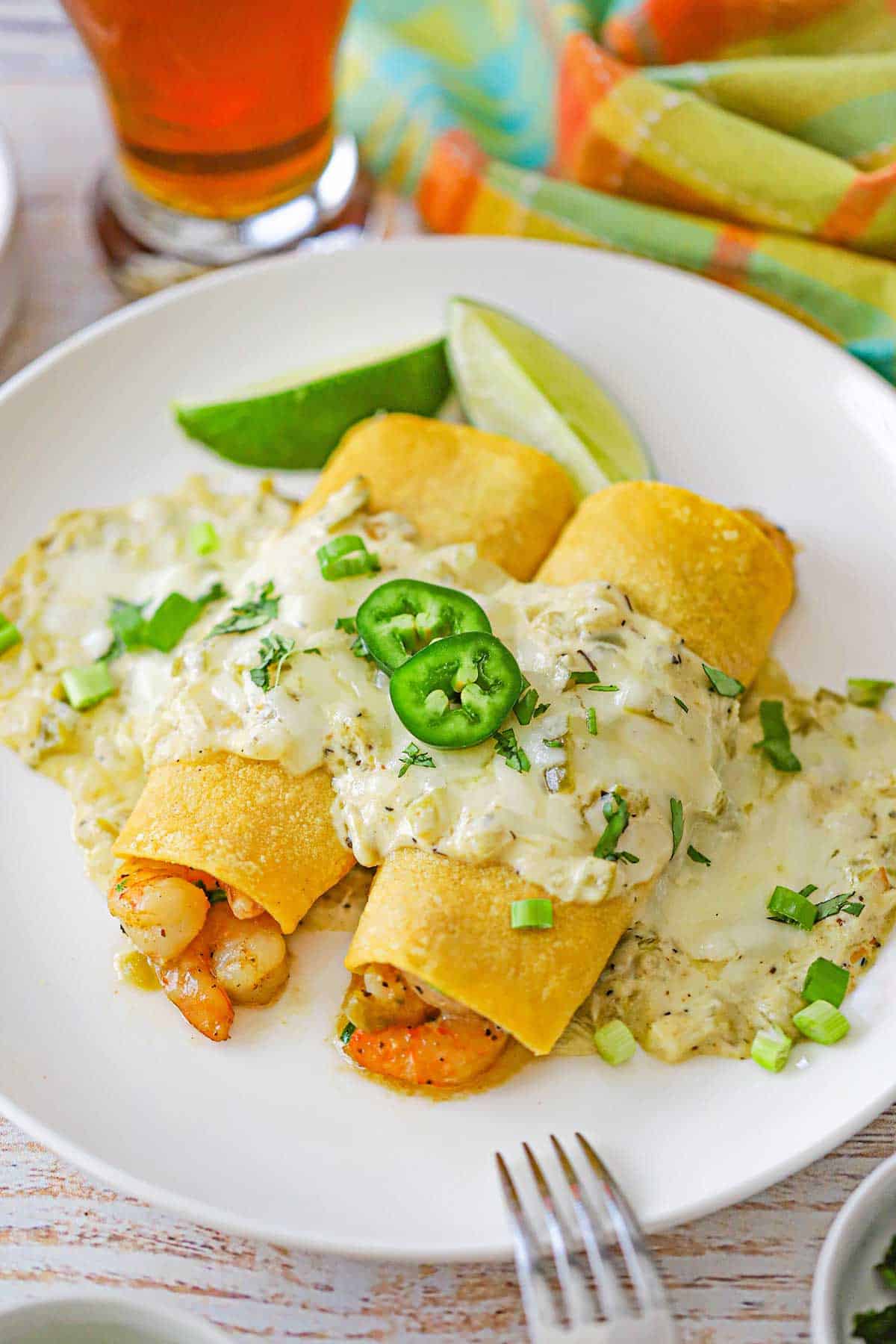 A white dinner plate filled with two creamy shrimp enchiladas that show the pink shrimp inside the rolled corn tortillas and topped with a creamy sauce and garnished with two sliced of jalapeños.