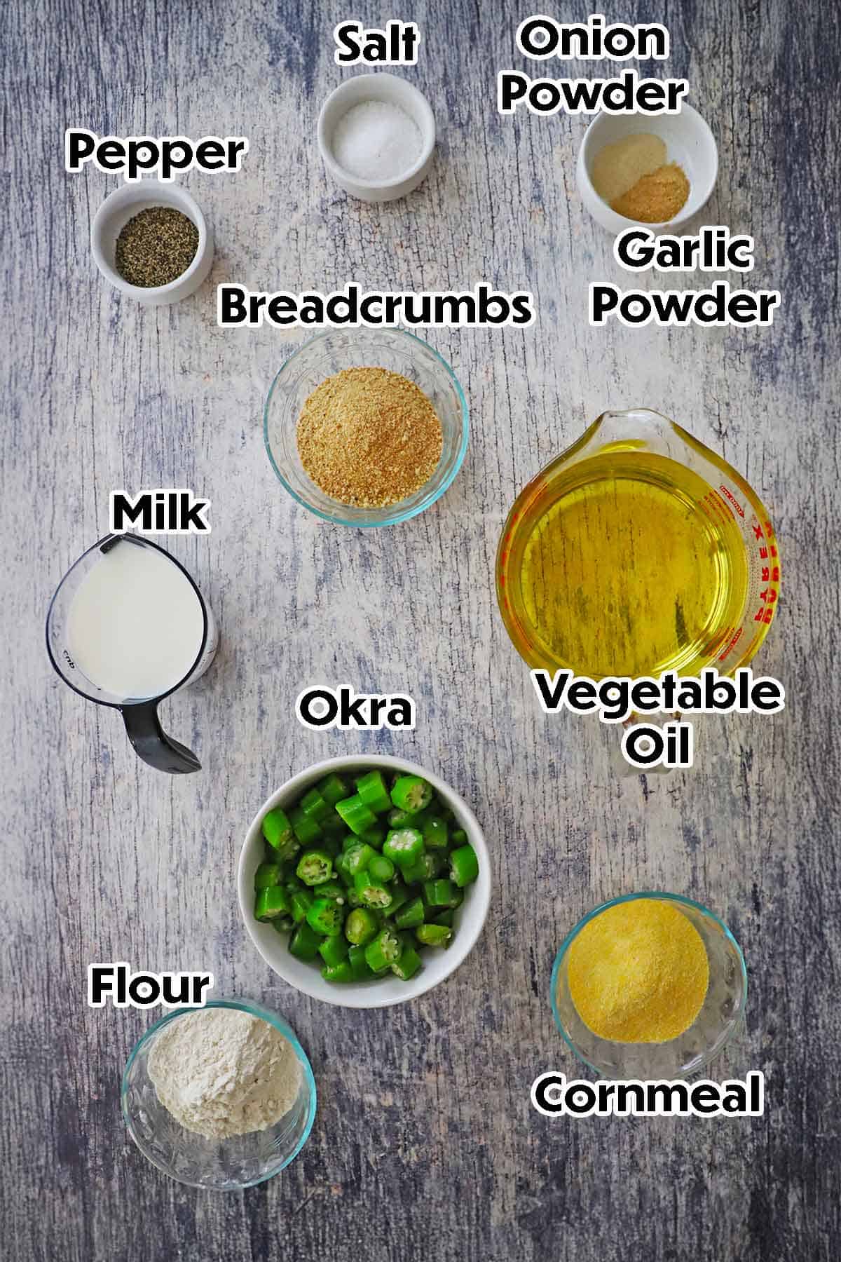 An arrangement of ingredients for Classic Southern Fried Okra on a grey wooden background including sliced okra, flour, cornmeal, milk, breadcrumbs, oil, and seasonings.