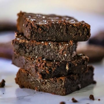 A close-up view of a stack of four ultimate fudge brownies with coarse sea salt sprinkled on the top of each brownie.