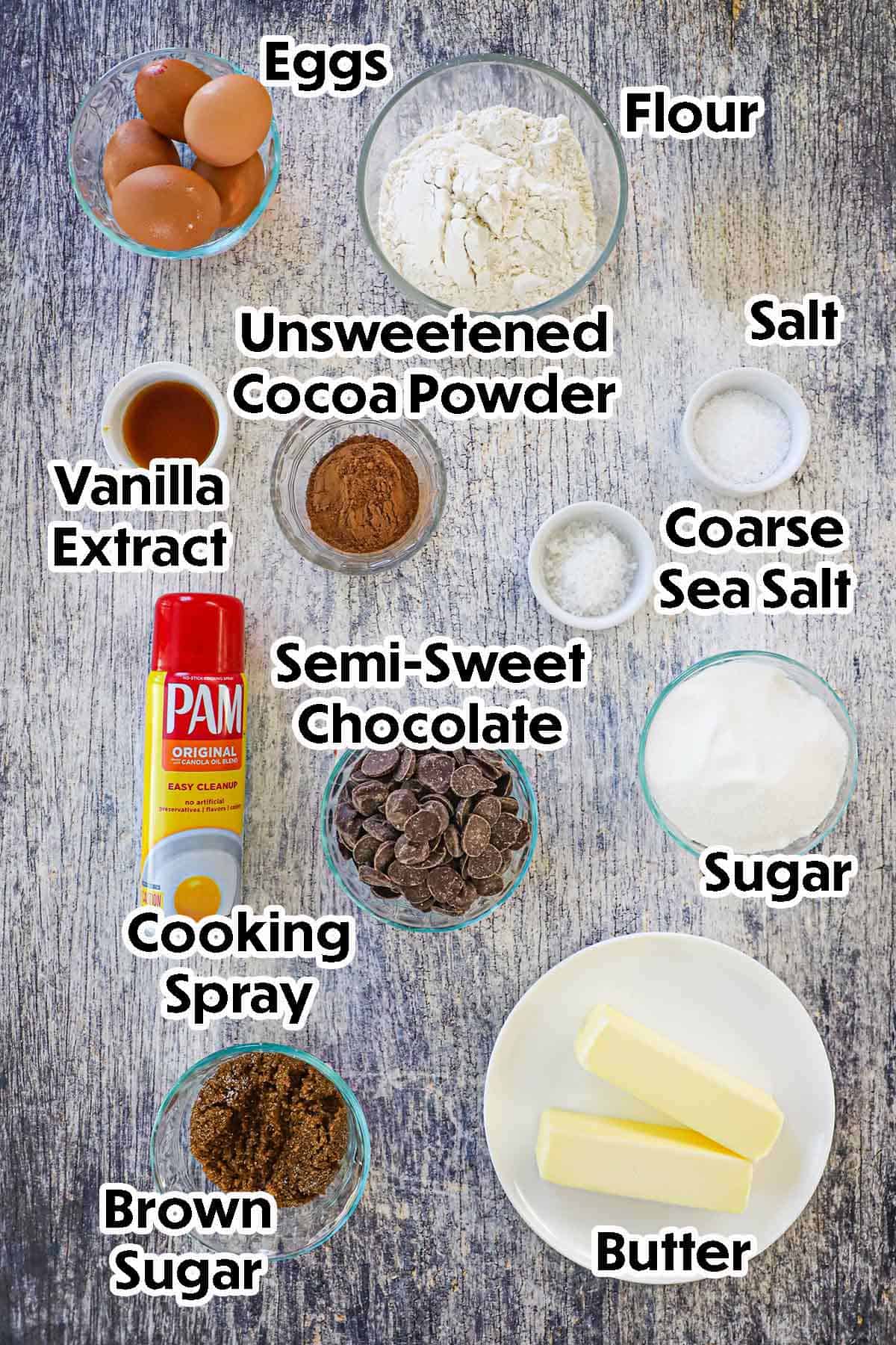 An arrangement of ingredients for fudge brownies on a grey wooden background including semi-sweet chocolate, butter, brown sugar, granulated sugar, eggs, cocoa powder, vanilla extract, salt, and cooking spray.