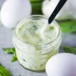A close-up view of a small jar filled with garlic basil aioli with a small spoon inserted into it with a couple of whole eggs and fresh basil resting nearby.