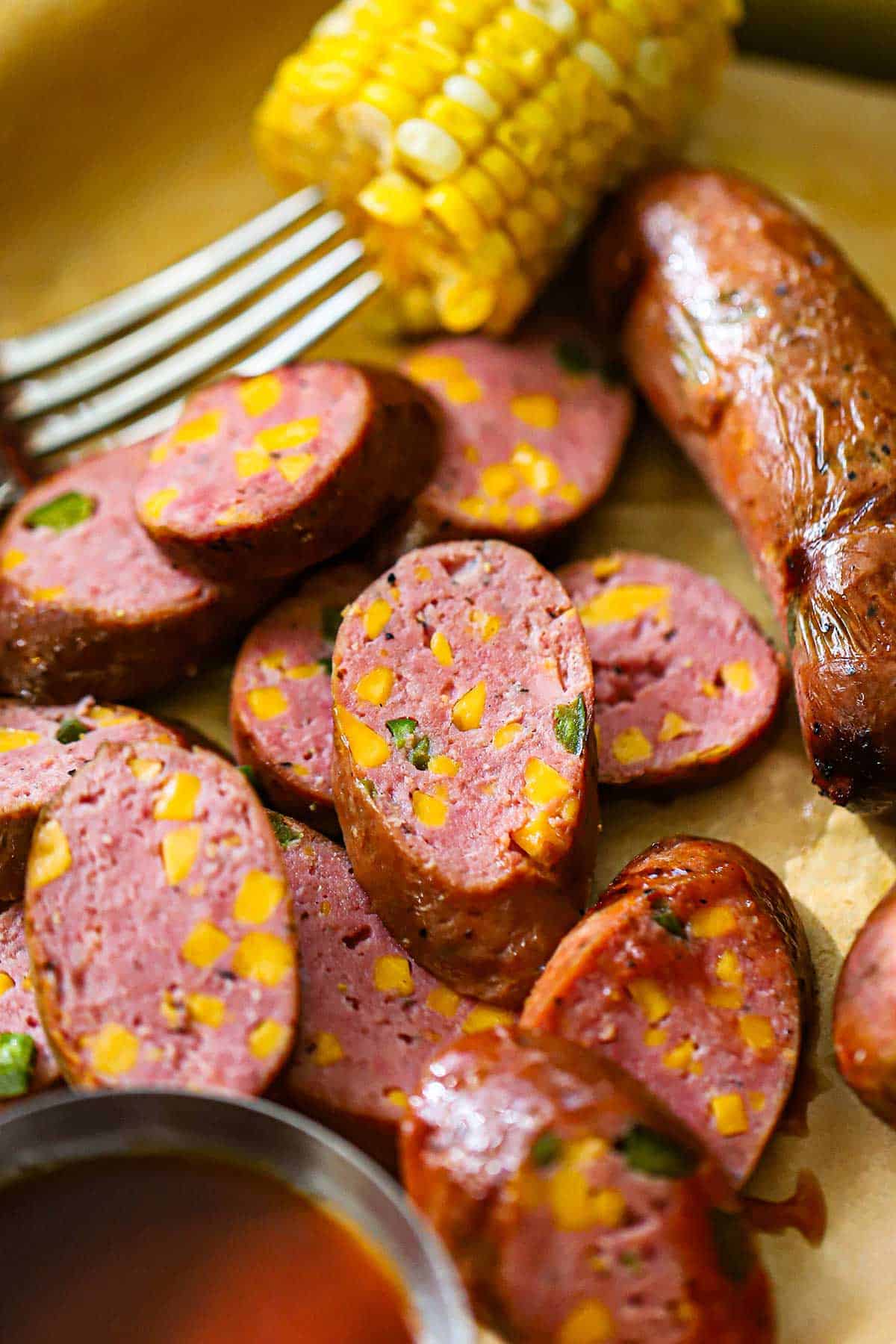 A close-up view of sliced Homemade Texas Jalapeño Cheddar Sausage on a tray lined with brown butcher paper and a corn on the cob niblet and small bowl of BBQ sauce nearby.