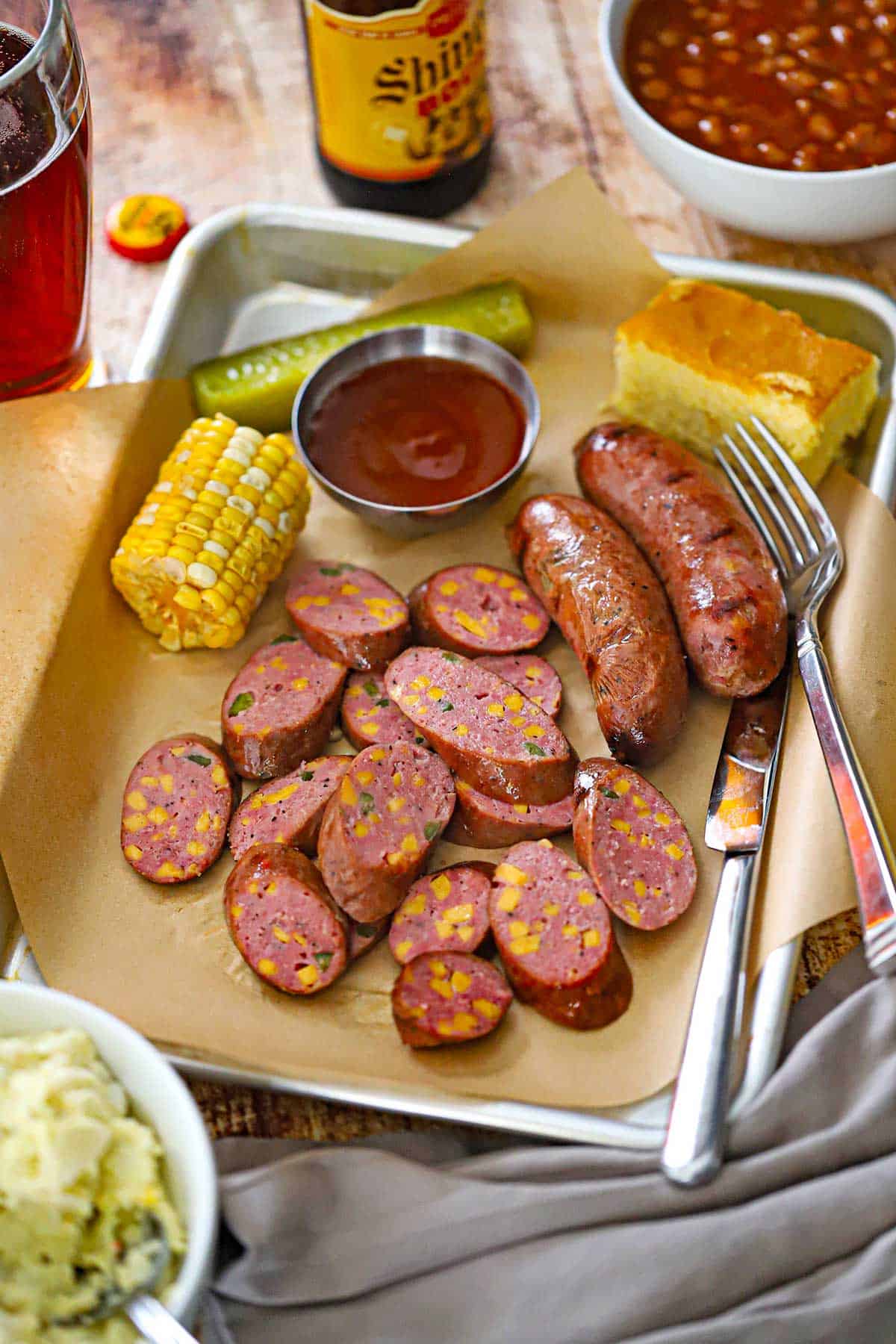 A metal trey lined with brown butcher paper filled with a serving of sliced Homemade Jalapeño Cheddar Sausage with a piece of corn on the cob, BBQ sauce, a pickle spear, and a square of corn bread all surrounding the sausage.