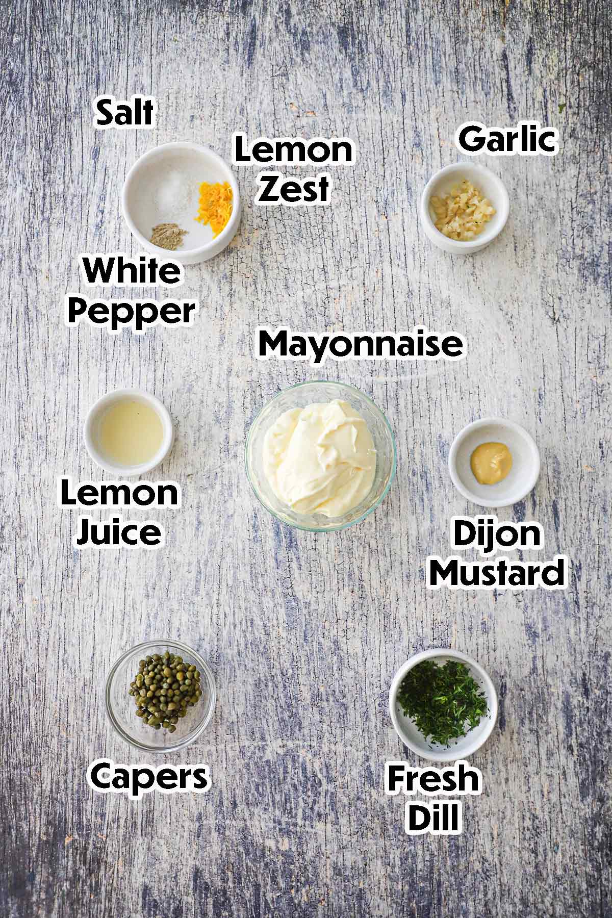 An arrangement of ingredients for Lemon-Dill and Caper Aioli on a grey wooden background including mayonnaise, Dijon mustard, capers, lemon juice, garlic, fresh herbs, and seasonings.