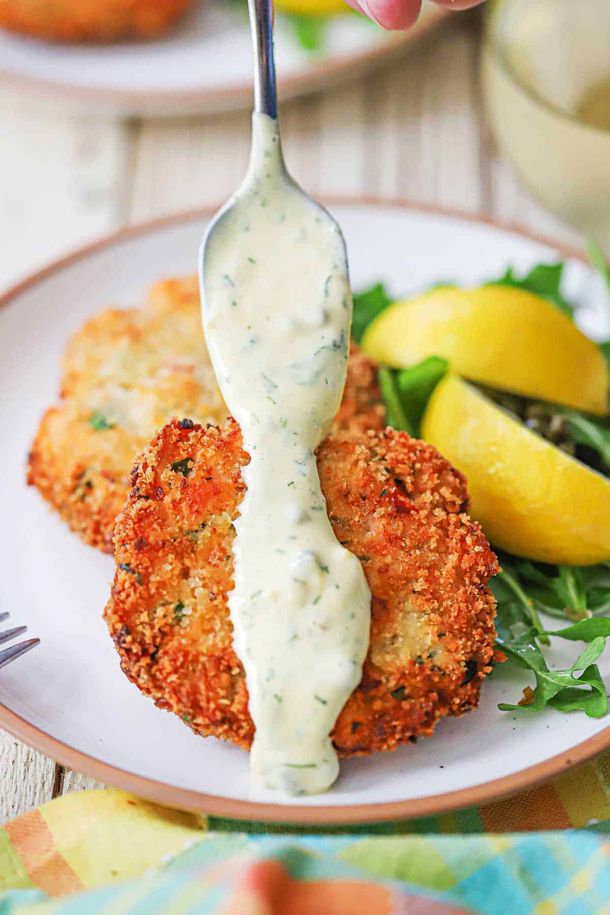 A person using a spoon to drizzle Lemon-Dill and Caper Aioli over the tops of two fresh salmon cakes nest to a couple of lemon wedges and arugula on a white plate.