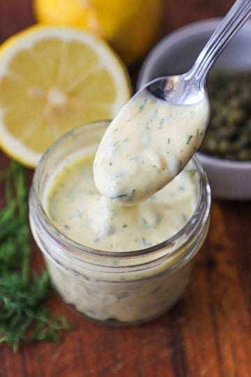 A spoon being used to hold up a serving of Lemon-Dill and Caper Aioli over a small jar containing the same with half of a lemon, dill sprigs, and capers nearby.