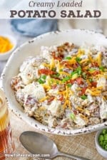 A side-view of a white serving bowl filled with a homemade creamy loaded potato salad topped with crispy bacon bits, chopped scallions, and shredded cheddar cheese.