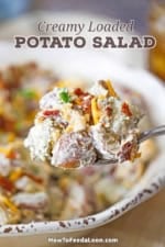 A large serving spoon holding a heaping mound of creamy loaded potato salad over a bowl of the same.