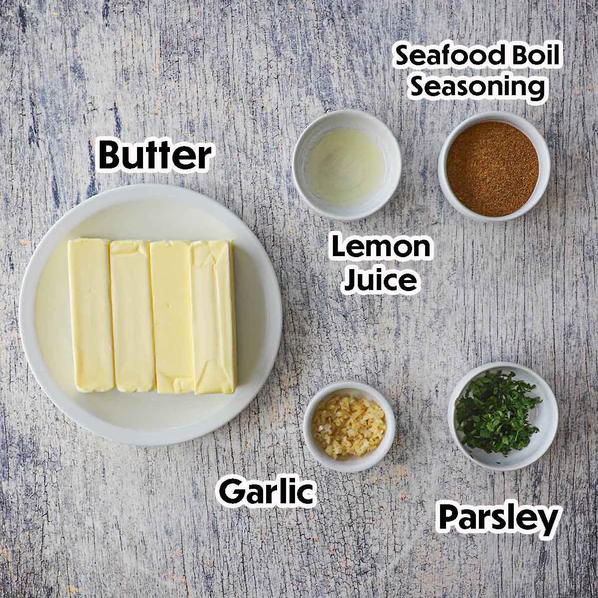 An arrangement of ingredients for Cajun Garlic Butter on a grey wooden background including four sticks of butter, lemon juice, garlic, Italian parsley, and seafood boil seasoning.