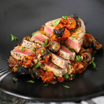 A close-up view of sliced Mediterranean tuna steak on black dinner plate topped with a black olive, tomatoes, and capers sauce and garnished with chopped Italian parsley.