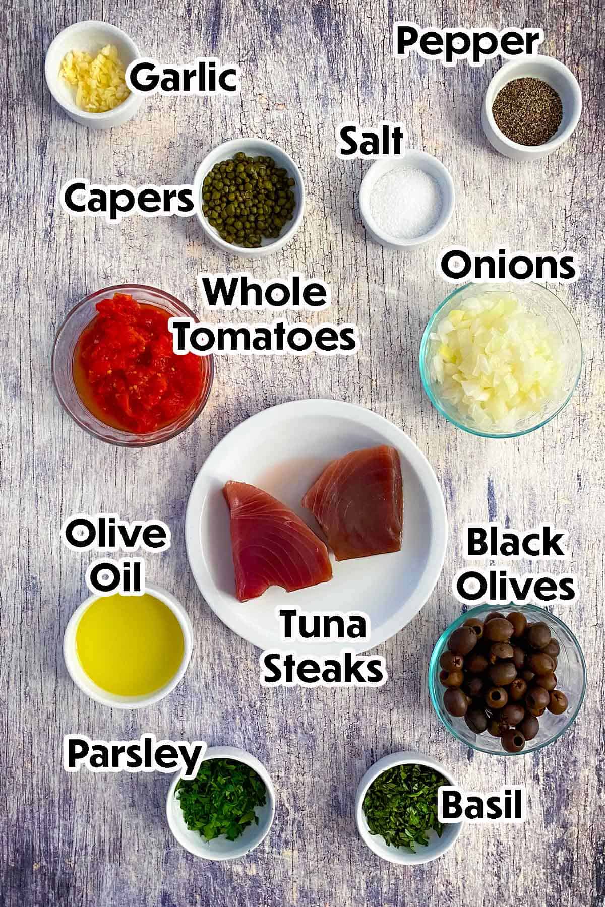 An arrangement of ingredients for Mediterranean Tuna Steaks on a grey wooden background including raw tuna steaks, tomatoes, onions, black olives, garlic, capers, herbs, and seasonings.