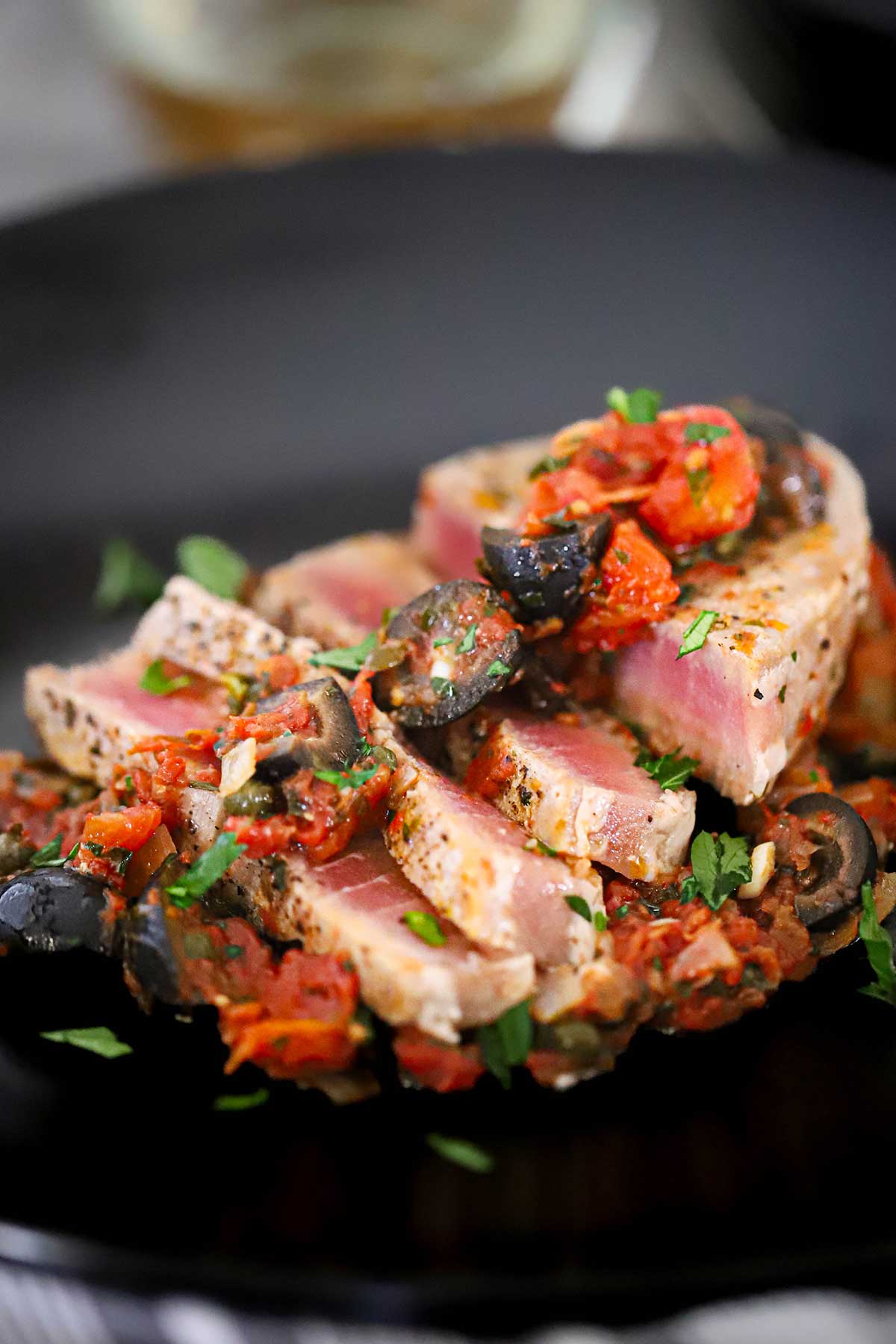 A close-up view of sliced Mediterranean tuna steak on black dinner plate topped with a black olive, tomatoes, and capers sauce.