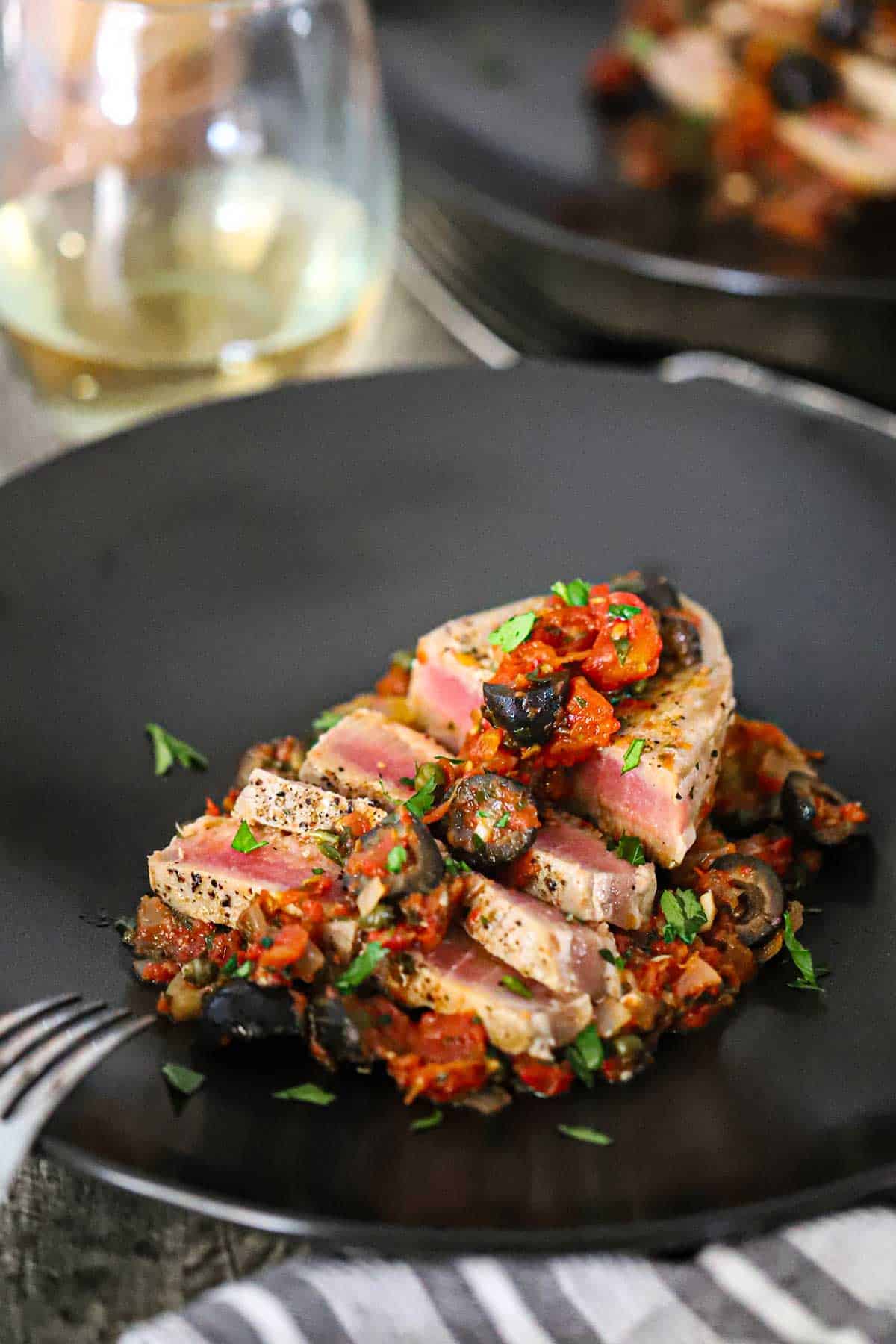 A straight-on view of sliced Mediterranean tuna steak on black dinner plate topped with a black olive, tomatoes, and capers sauce.