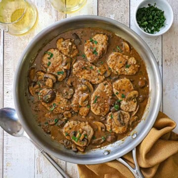 An overhead view of a large stainless steel skillet filled with Pork Medallions with Mushroom Gravy with a serving spoon, two glasses of wine, and a small bowl of chopped parsley all nearby.