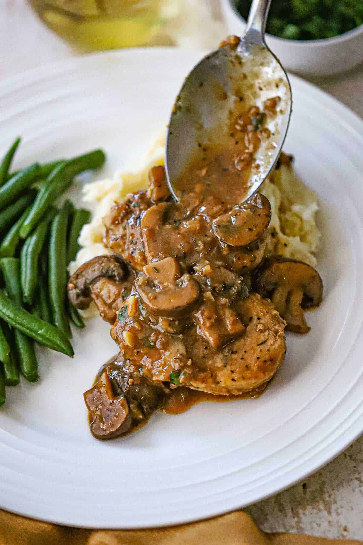 A person using a serving spoon the drizzle a Dijon mushroom gravy sauce over the top of two pan-seared pork medallions on a white plate with mashed potatoes and green beans.