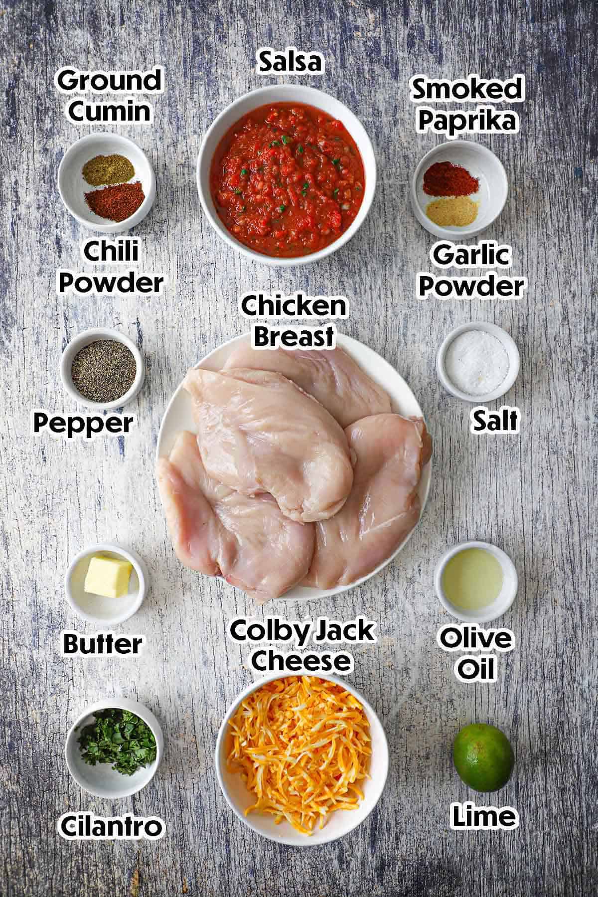 An arrangement of ingredients for Easy Cheesy Salsa Chicken on a grey wooden background including raw chicken breasts, salsa, shredded cheese, cilantro, butter, oil, and seasonings.