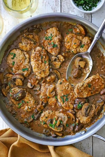 An overhead view of a large stainless steel skillet filled with Pork Medallions with Mushroom Gravy two glasses of wine, and a small bowl of chopped parsley all nearby and a serving spoon in the sauce in the skillet.