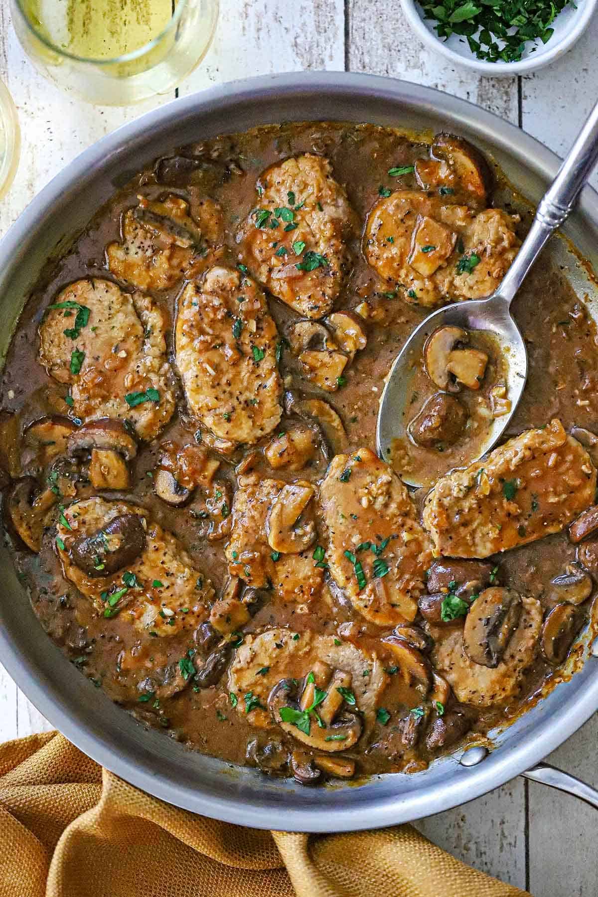 An overhead view of a large stainless steel skillet filled with Pork Medallions with Mushroom Gravy two glasses of wine, and a small bowl of chopped parsley all nearby and a serving spoon in the sauce in the skillet.