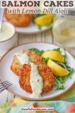 Two fresh salmon cakes topped with a streak of lemon-dill and caper aioli next to two lemon wedges on a white plate.