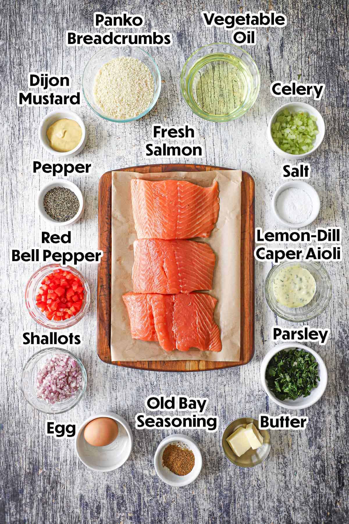 An arrangement of ingredients for Fresh Salmon Cakes with Lemon-Dill Aioli on a grey wooden background including fresh salmon, breadcrumbs, bell peppers, shallots, an egg, butter, celery, Dijon mustard, oil, aioli, herbs, and seasonings.