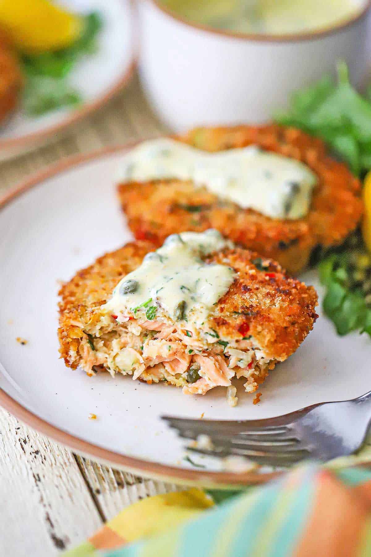 A close-up view of a half-eaten Salmon Cake topped with a smear of Lemon-Dill Aioli on a white plate with another salmon cake in the background.