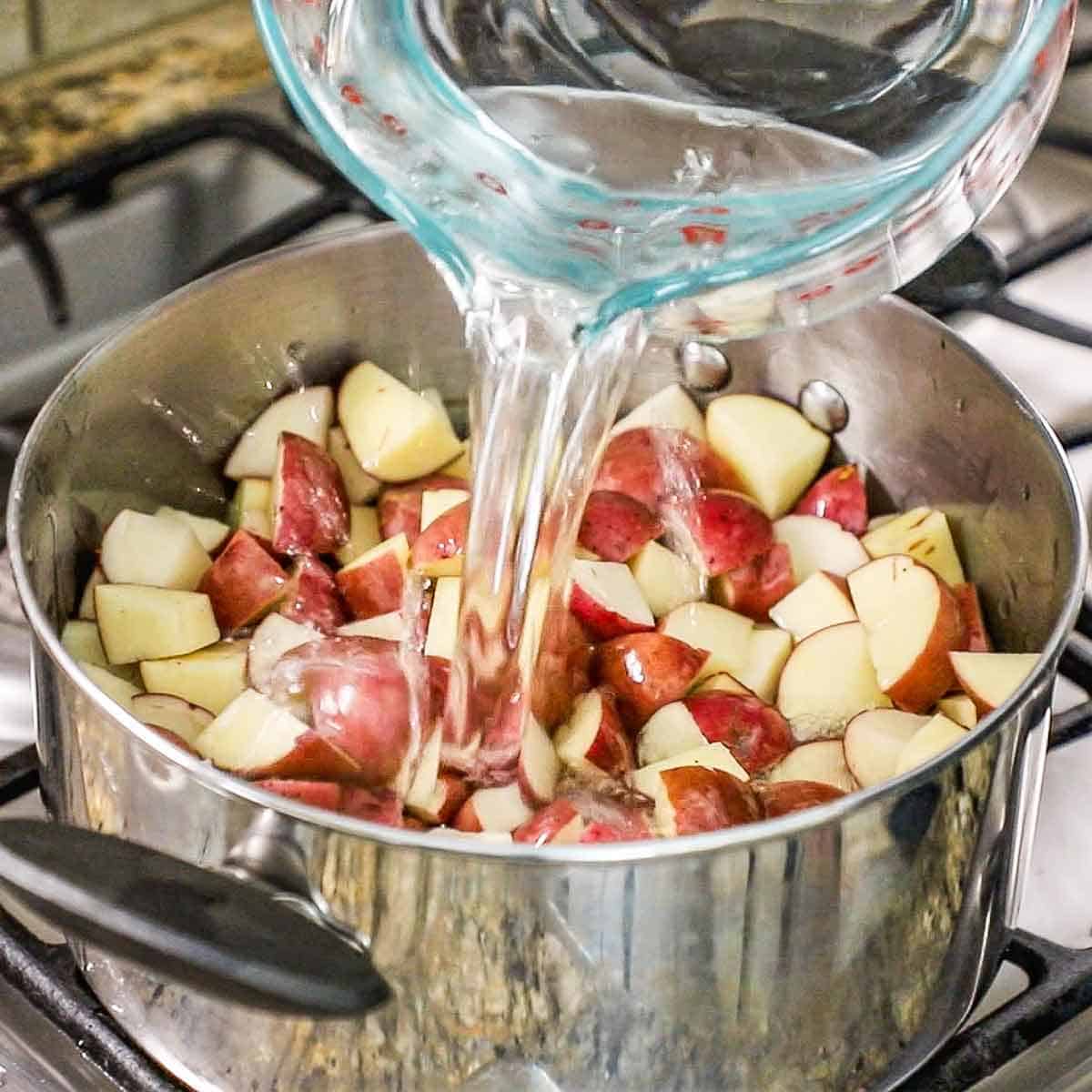 A person pouring water from a large glass measuring cup into a pot on a gas stove that is filled with cubed red potatoes.
