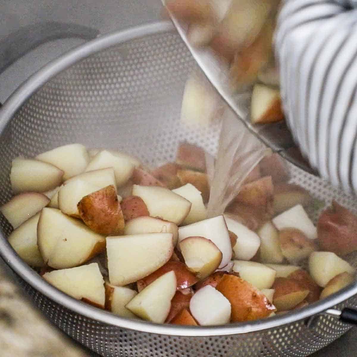 A person dumping cooked red potatoes and boiling water from a pot into a colander in a sink.