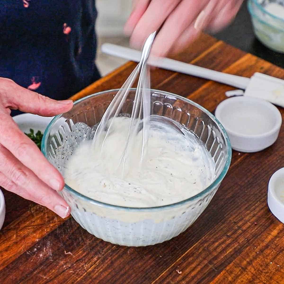 A person using a whisk to combine sour cream, mayonnaise, and apple sider vinegar in a glass bowl.