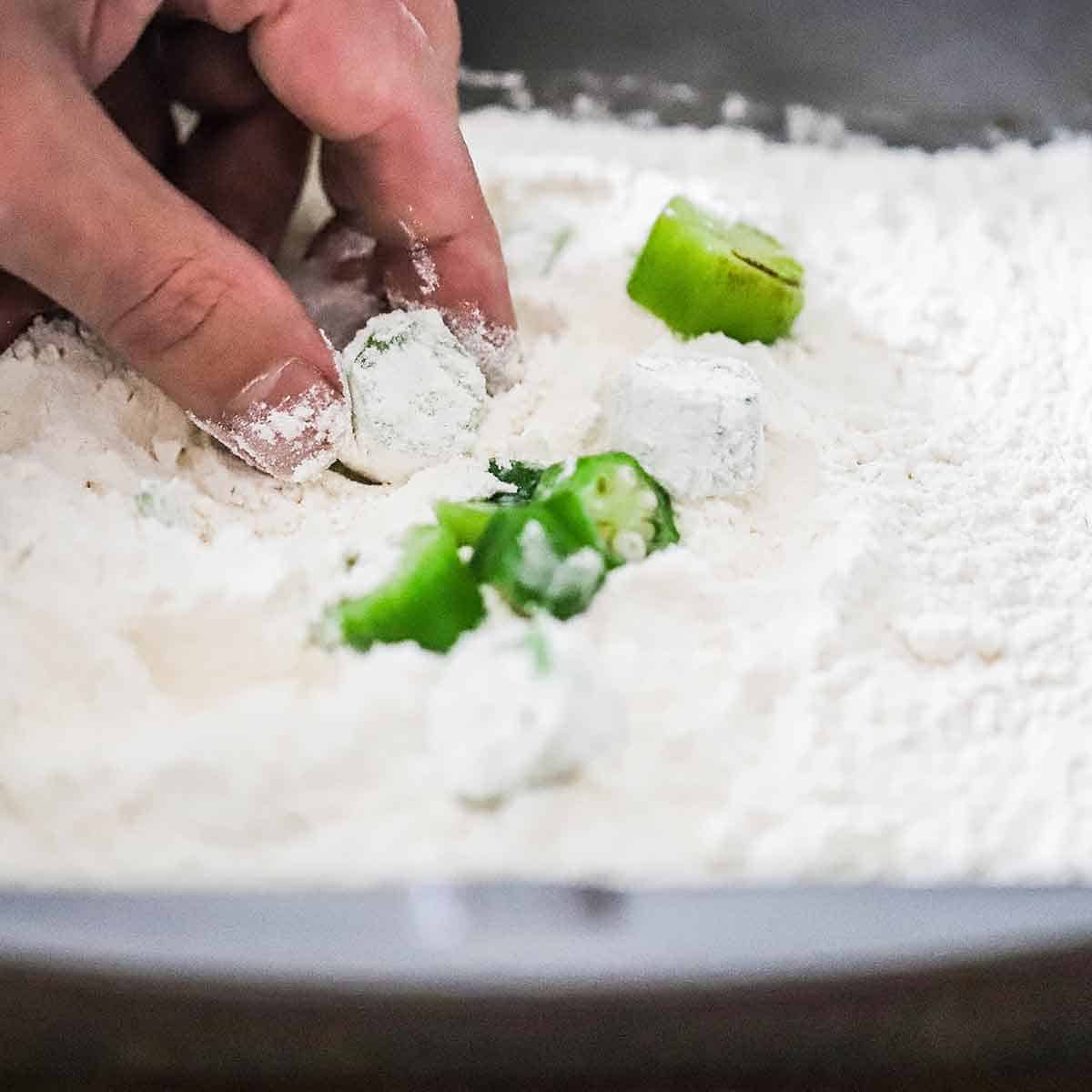 A person using his fingers to coat sliced okra in a pan that is filled with all-purpose flour.