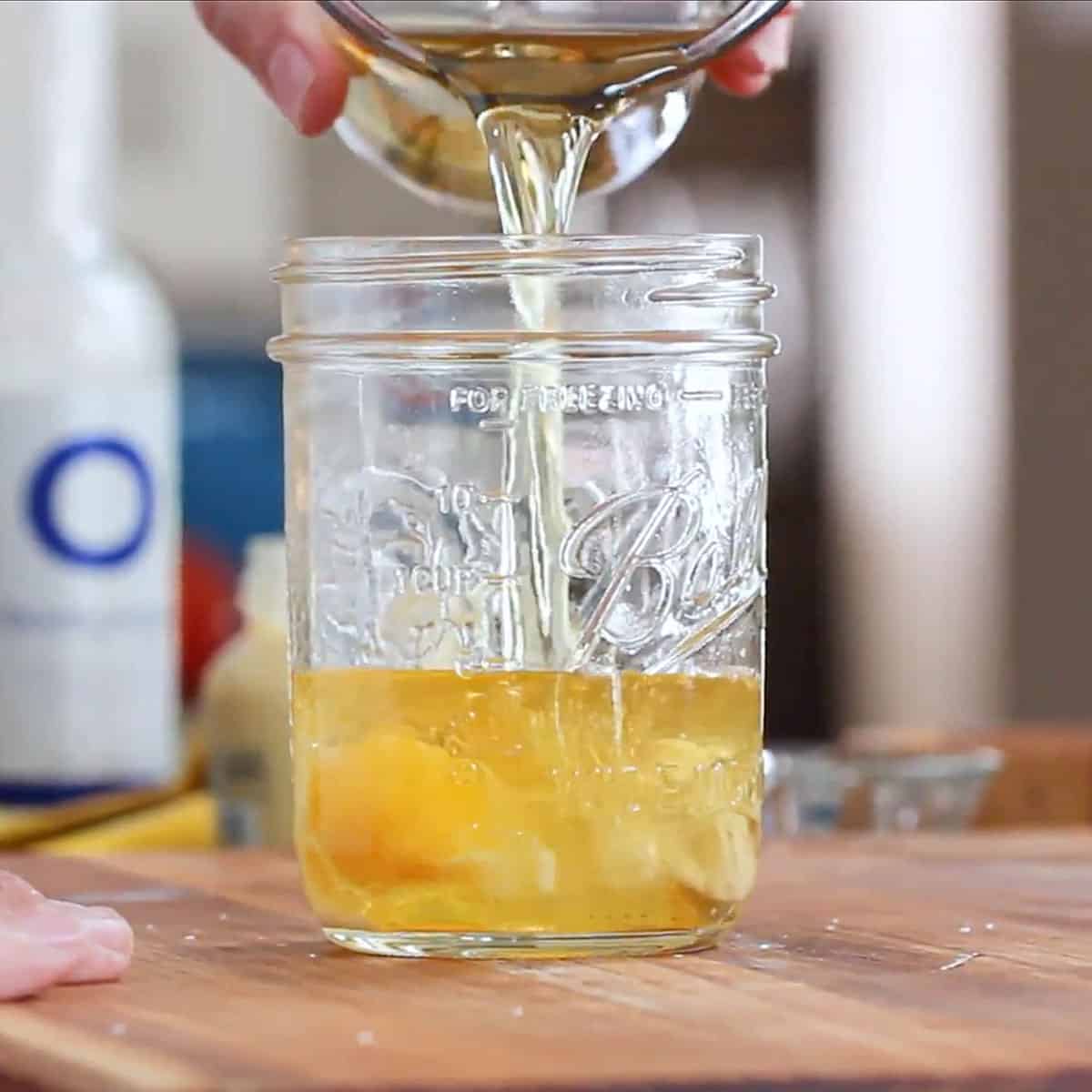 A person pouring avocado oil into a jar that is filled with an egg yolk, a whole egg, white wine vinegar, and lemon juice.