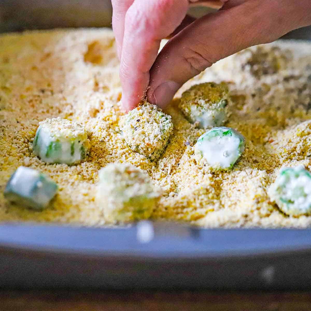 A person dredging pieces of okra that have been coated with flour and milk through breadcrumbs in a baking pan.