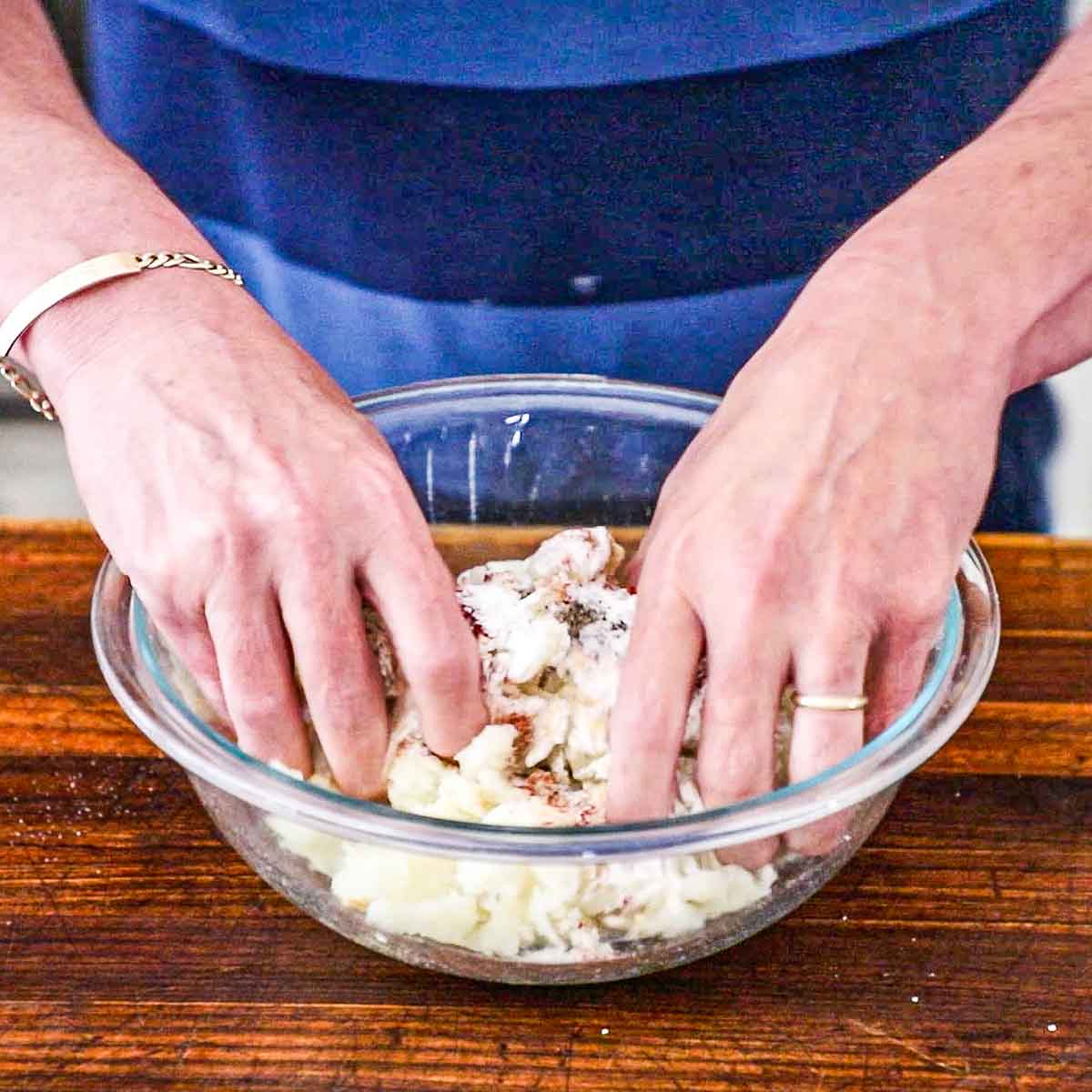 A person using both of his hands to mix shredded par-boiled Russet potatoes in a glass bowl with seasonings.