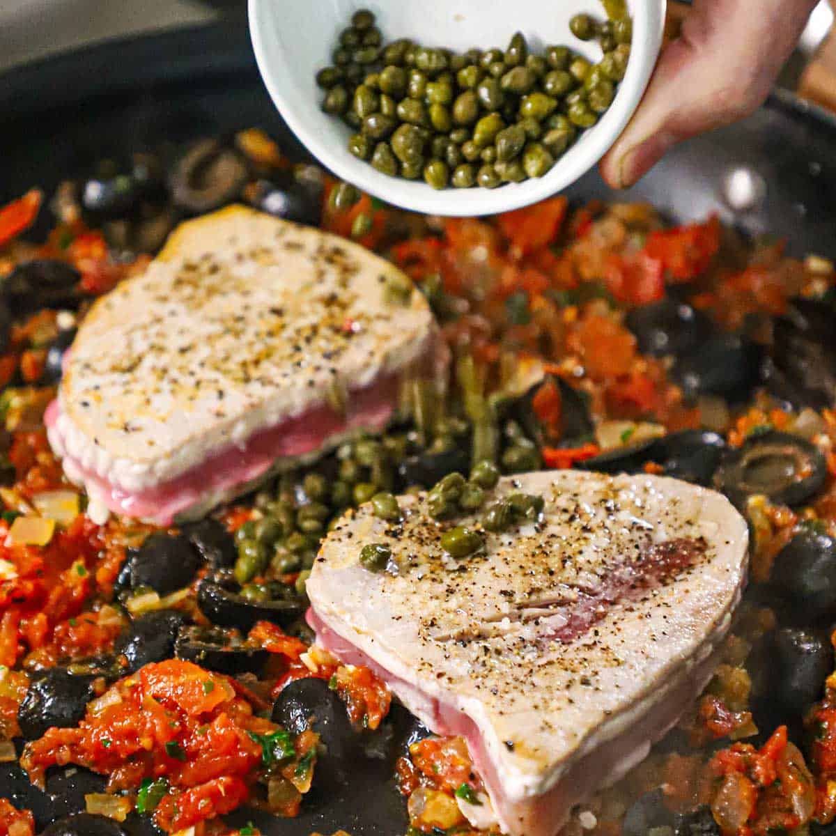 A person adding capers to a skillet fillet with two Mediterranean tuna steaks simmering in a tomato and black olive sauce.