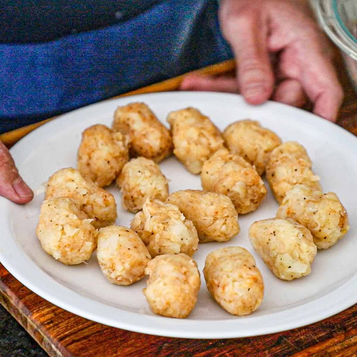 A person holding a white dinner plate that is filled with formed homemade tater tots before they have been fried.