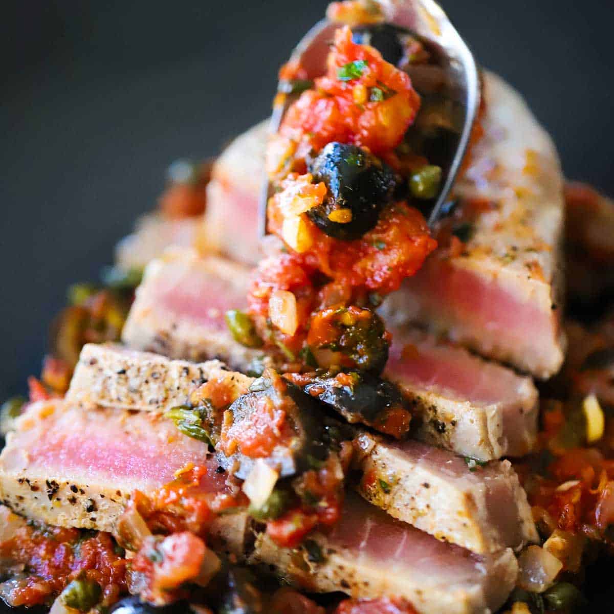A person using a spoon to spread a line of a tomato, black olive, and caper sauce over the top of a sliced pan-seared tuna steak.