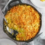 An overhead view of a large cast-iron skillet filled with a serving of summer squash casserole with a portion of it missing with a serving spoon in the pan.