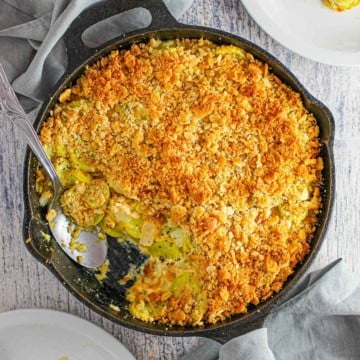 An overhead view of a large cast-iron skillet filled with a serving of summer squash casserole with a portion of it missing with a serving spoon in the pan.