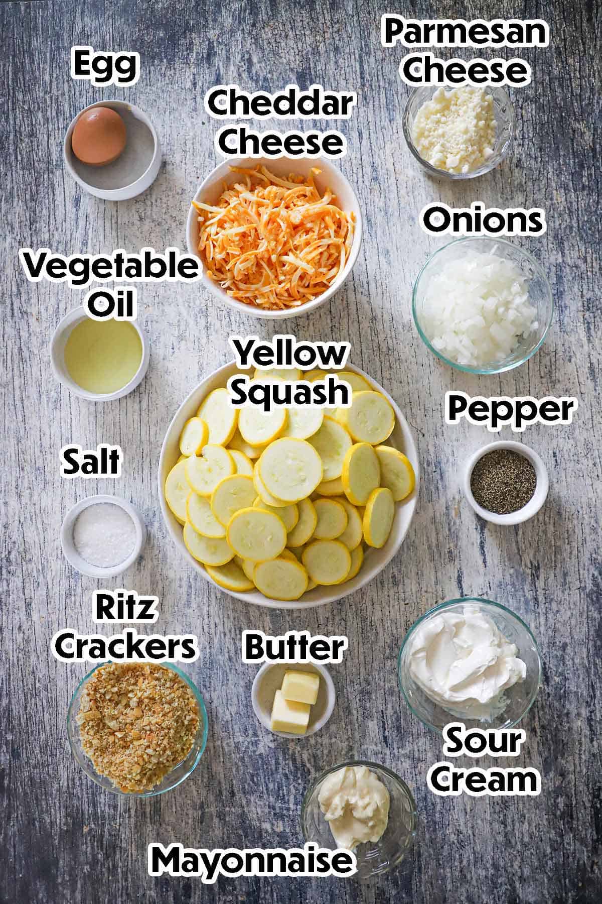 An arrangement of ingredients for Summer Squash Casserole on a grey wooden background including sliced yellow squash, onions, cheddar cheese, an egg, Parmesan cheese, oil, sour cream, mayonnaise, butter, Ritz crackers, and seasonings.