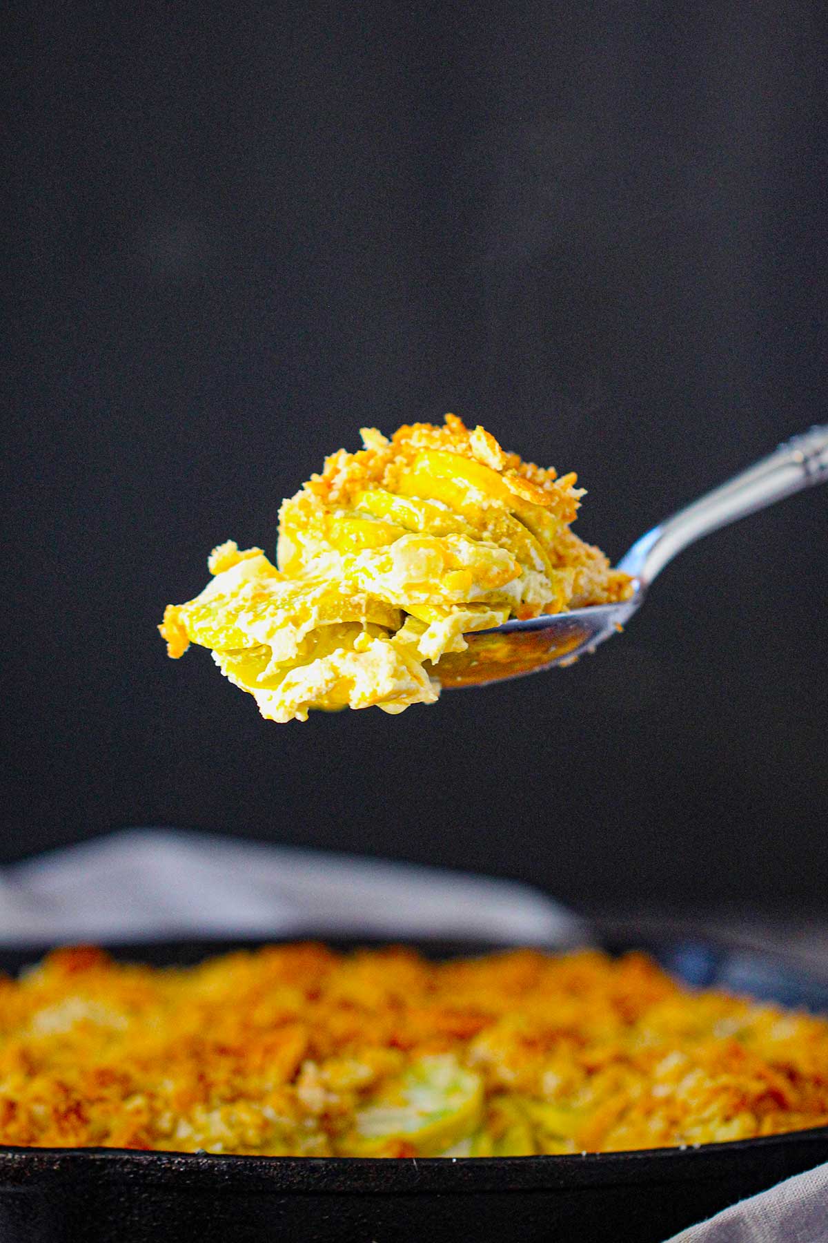 A person using a large serving spoon to lift a heaping helping of Summer Squash Casserole from a cast-iron skillet filled with the same.