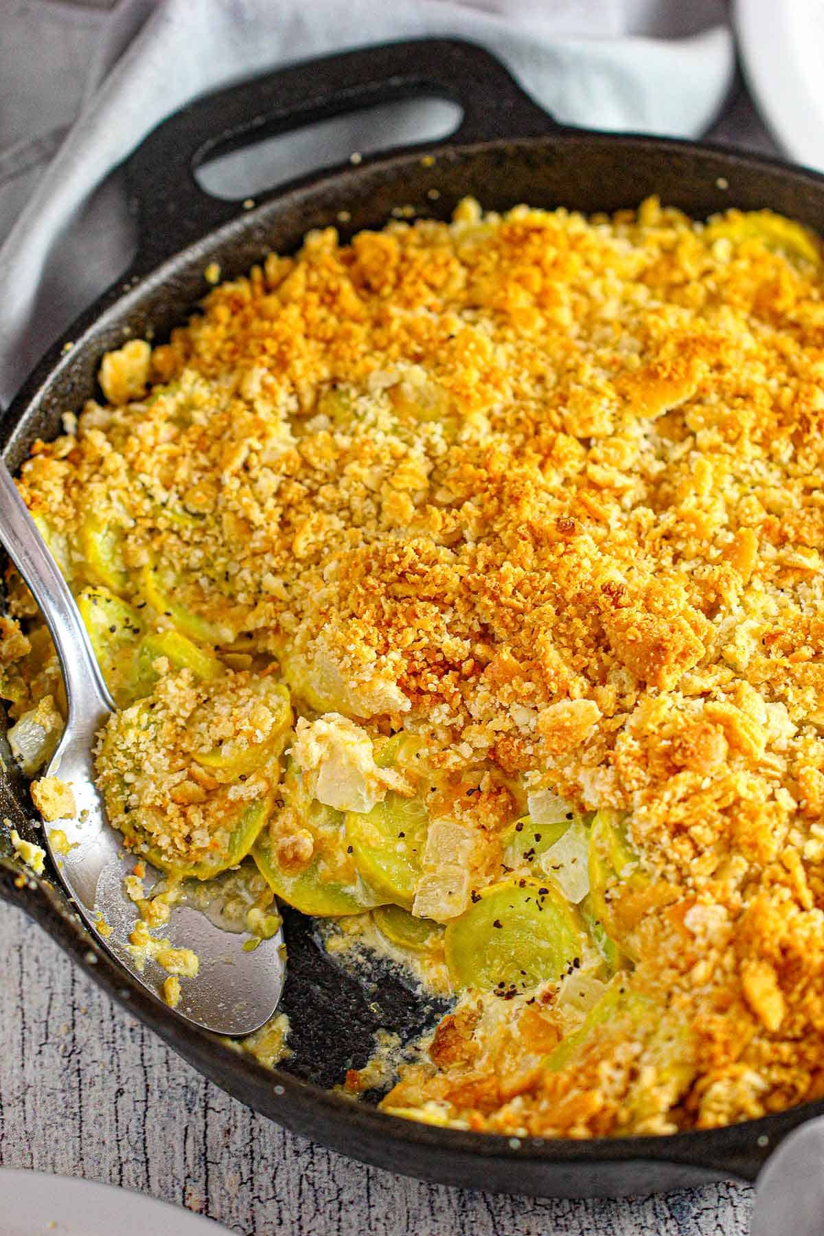 A close-up view of a large cast-iron skillet filled with a serving of summer squash casserole with a portion of it missing with a serving spoon in the pan.