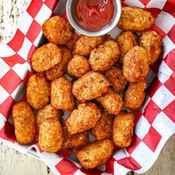 A close-up overhead view a basket lined with a red and white checkered paper and filled with crispy homemade tater tots with a small vessel of ketchup nearby.