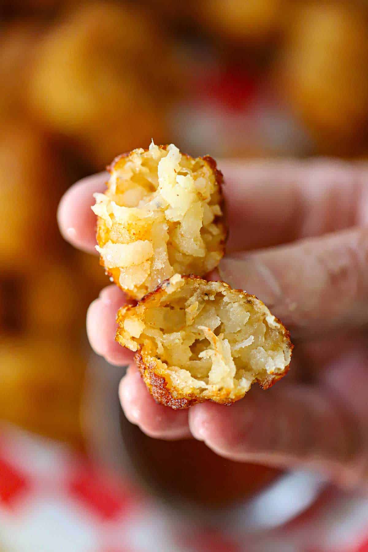 A person holding two halves of a homemade tater tot that has been split in half revealing the soft shreds of potato on the inside of the crispy tater tot.