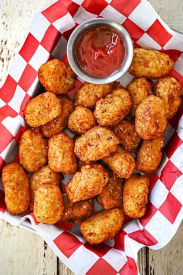 An overhead view a basket lined with a red and white checkered paper and filled with crispy homemade tater tots with a small vessel of ketchup nearby.