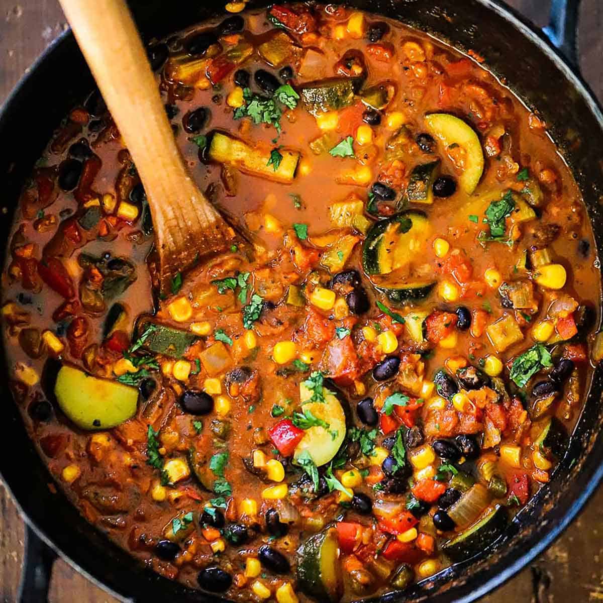 An close-up overhead view of a large black cast-iron pot filled with simmering Vegetable Chili with Black Beans on a gas stove.